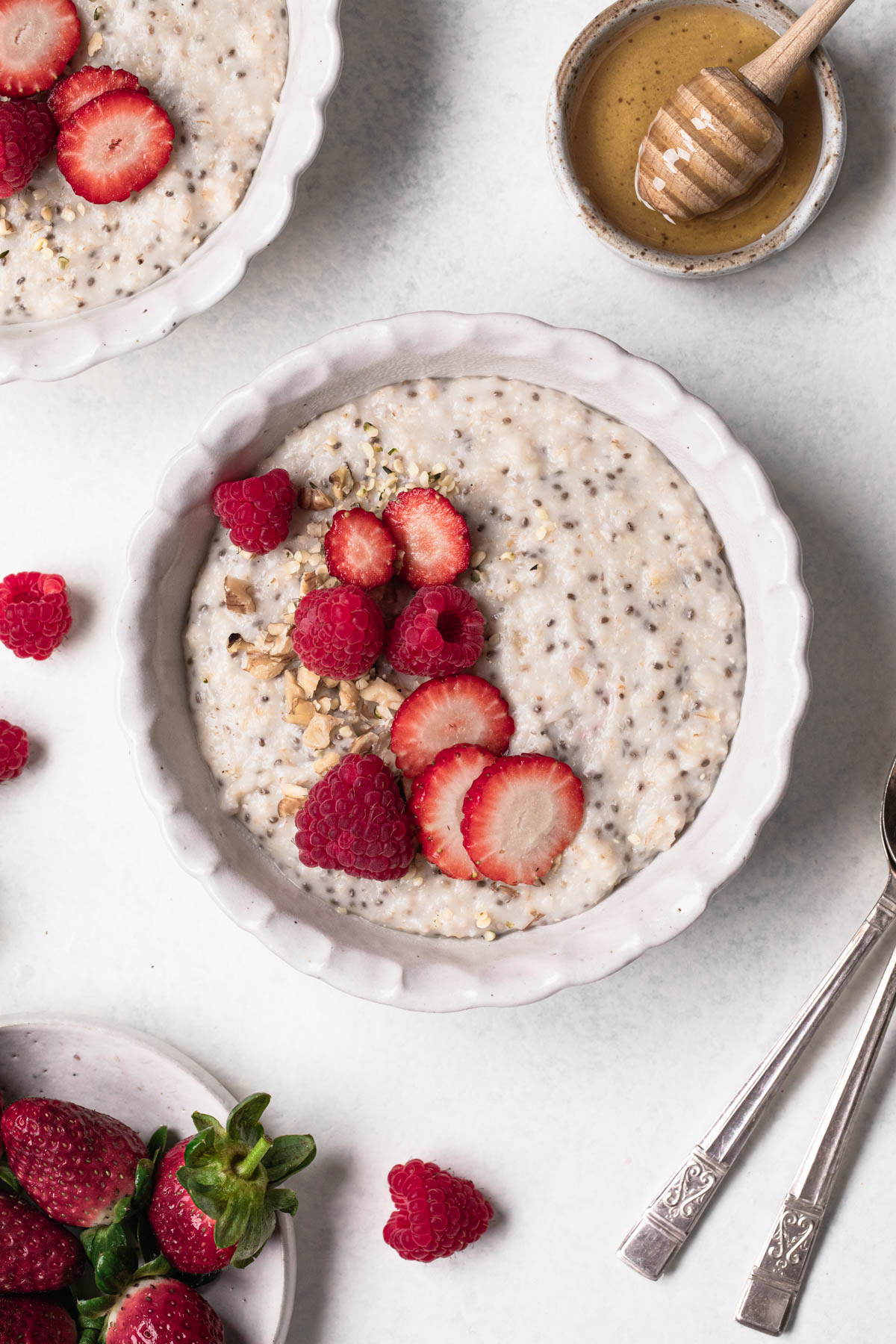 White ceramic bowl of porridge with chia seeds and sliced strawberries, on a white table with strawberries and honey