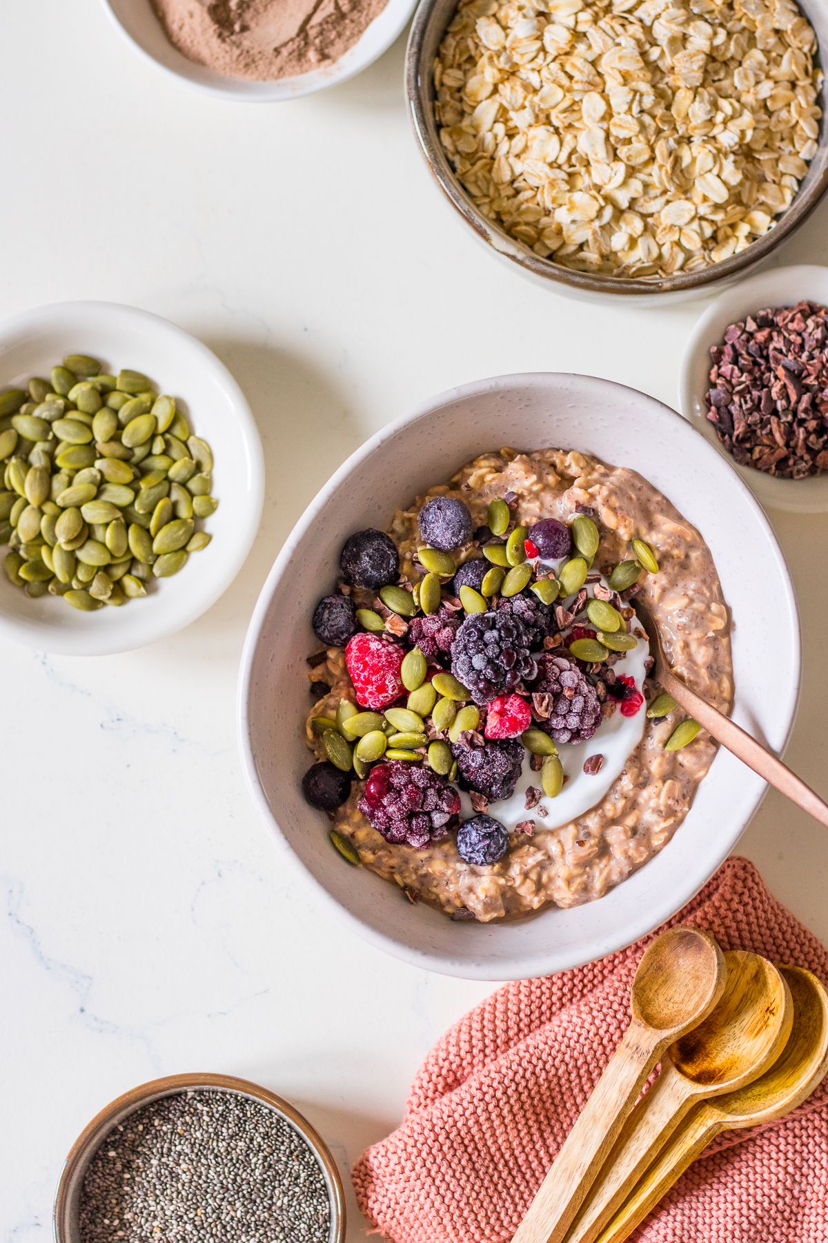 Chocolate oatmeal in a bowl with berries and pumpkin seeds, surrounded by small dishes of seeds and cacao nibs
