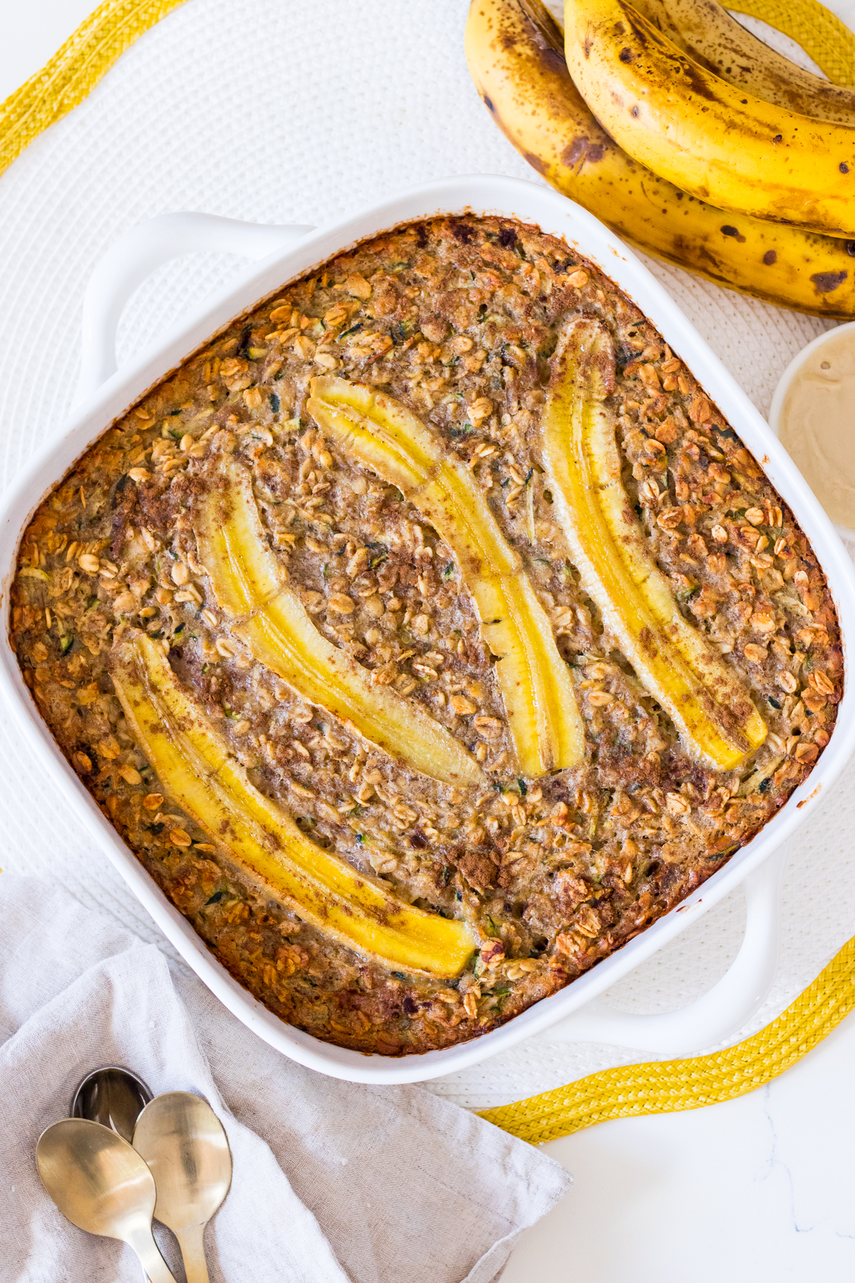 Top view of banana baked oats in a white ceramic dish with a serve of baked oats on a side plate with yoghurt