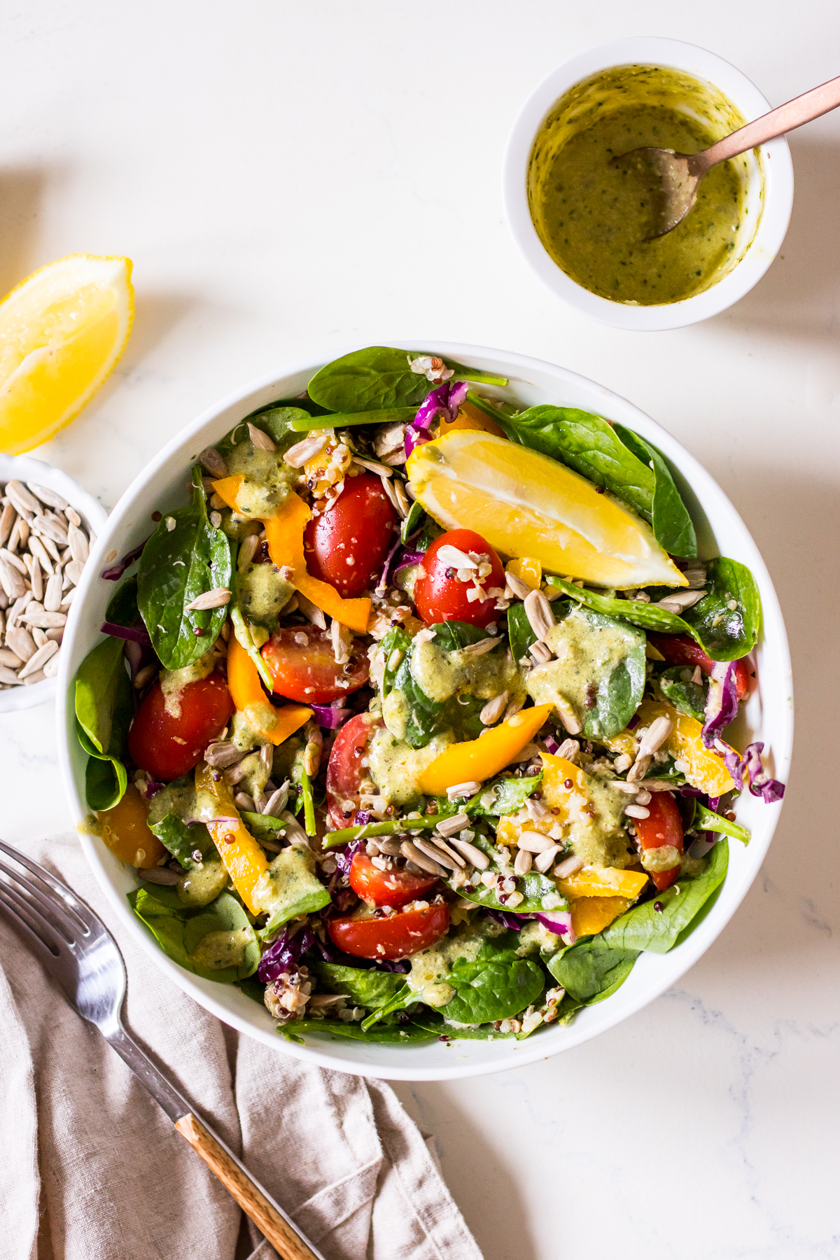 An image of a salad with tuna, spinach, tomatoes, quinoa and capsicum in a white bowl, with a pesto dressing to the side