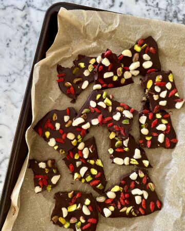 Dark chocolate bark topped with chopped pistachios, macadamias and goji berries, set on a baking tray lined with parchment paper