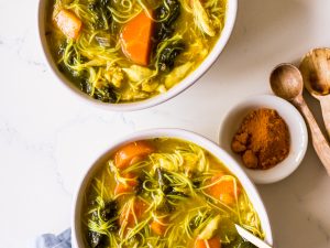 Two bowls of turmeric chicken noodle soup with gold spoons and a blue napkin