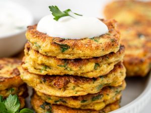 Stack of gluten free corn fritters on a white plate topped with yoghurt and herbs.